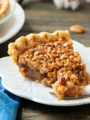 A slice of old fashioned pecan pie on a white plate with a fork. A pumpkin and a whole pie can be seen in the background.