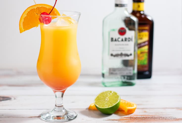 hurricane cajun cocktail with orange, cherry, and lime. 2 bottles of rum are seen in the background.