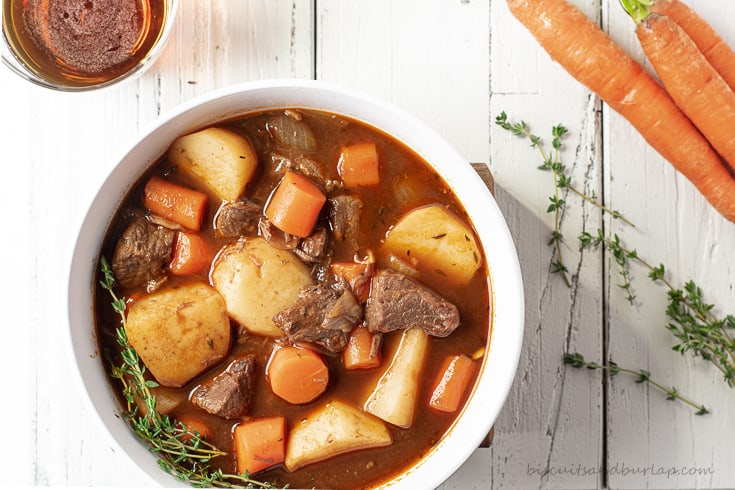 A white bowl of Guinness Irish stew with a glass of beer and carrots scattered nearby.