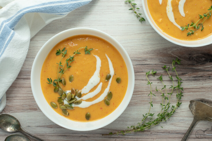 Red pepper and butternut squash soup in white bowls on a wooden table topped with pumpkin seeds and fresh thyme