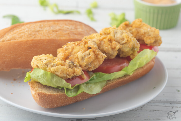 fried oyster po'boy on white plate with lettuc and tomatoes