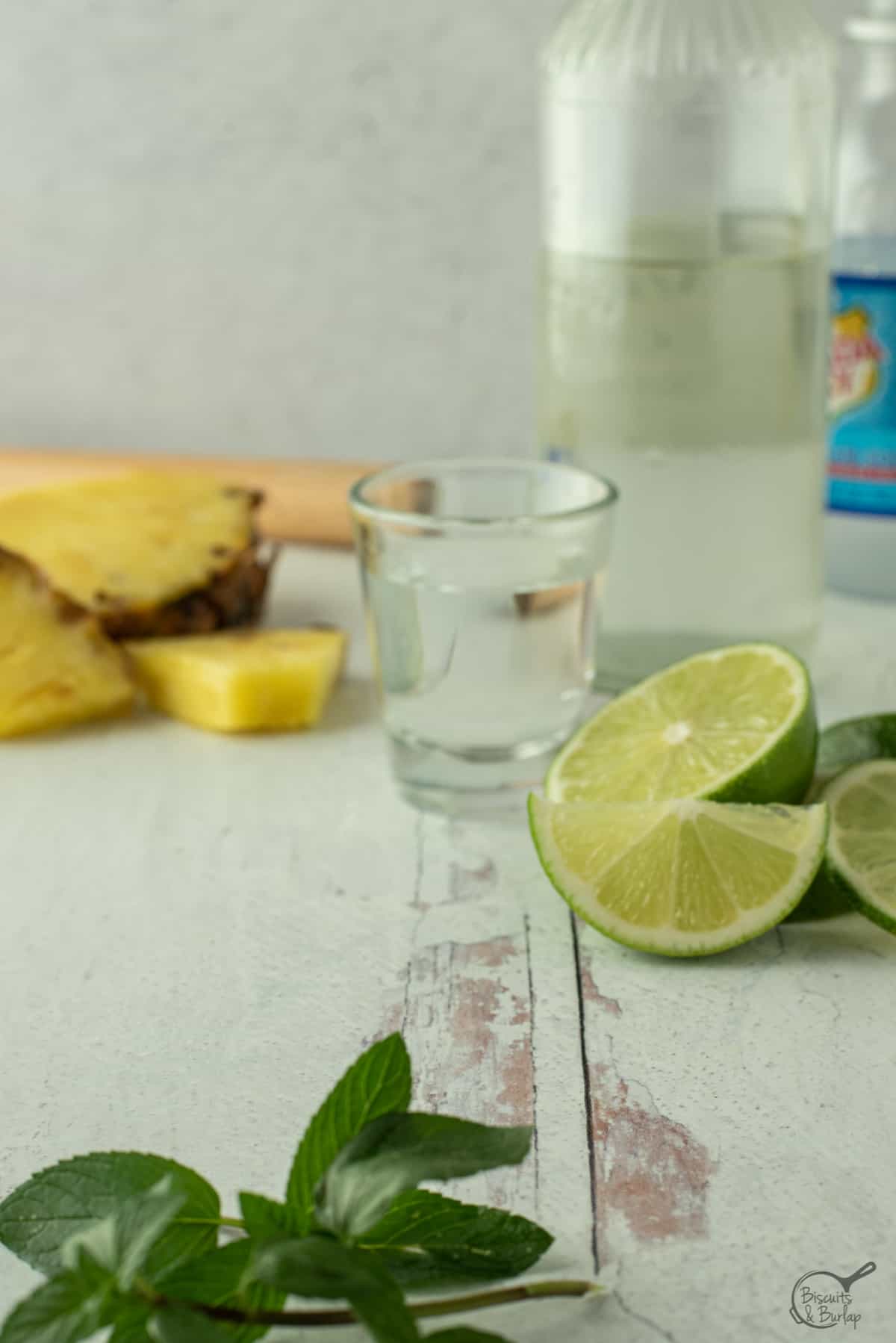 shot glass, lime slices, fresh pineapple, and mint leaves on white wood background