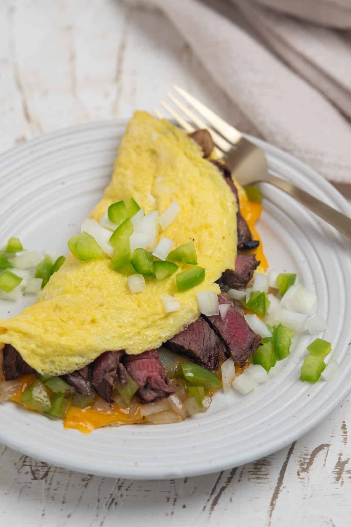 omelette stuffed with steak, cheese, peppers, onions, and mushrooms on a white plate