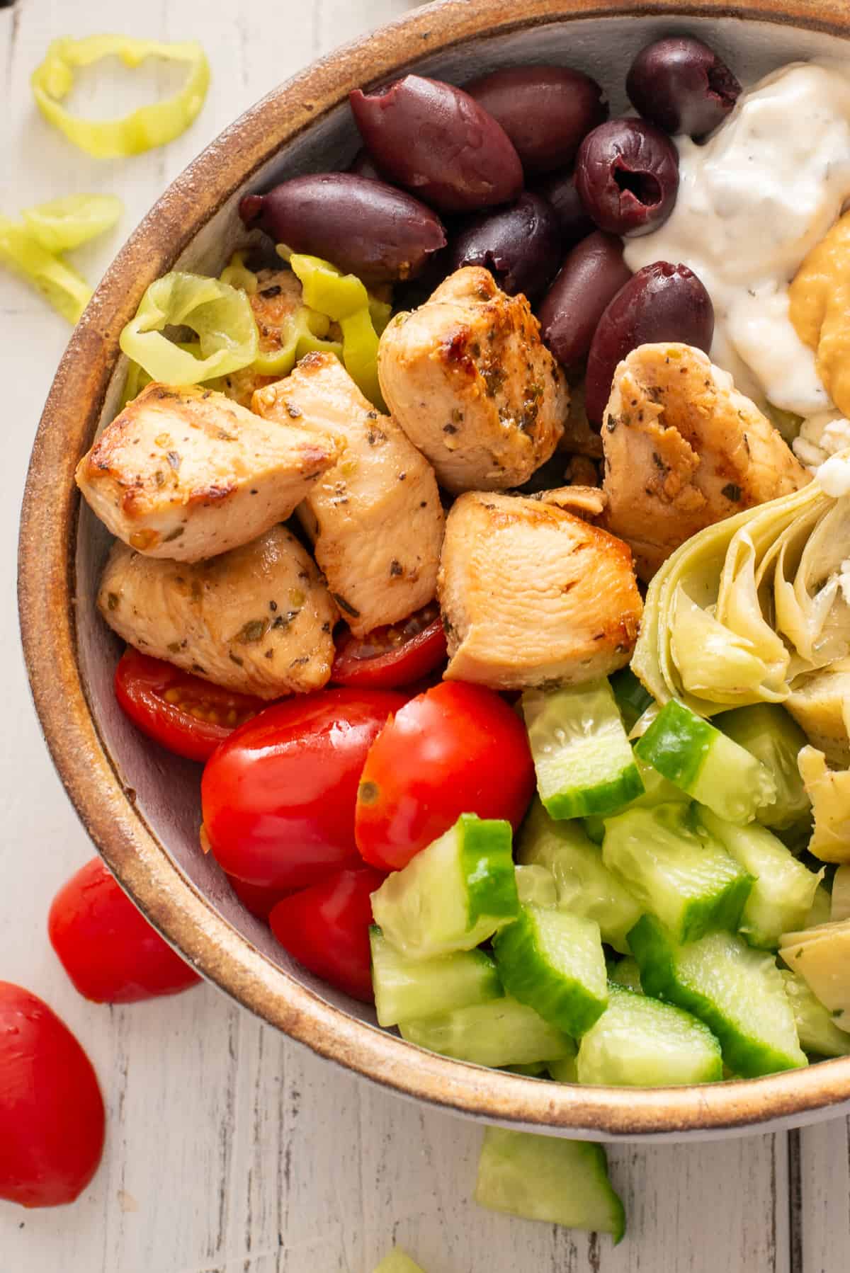 Close up of Greek chicken bowl with kalamata olives, pepperonicini peppers, chicken, tomatoes, cucumbers, and artichoke hearts.