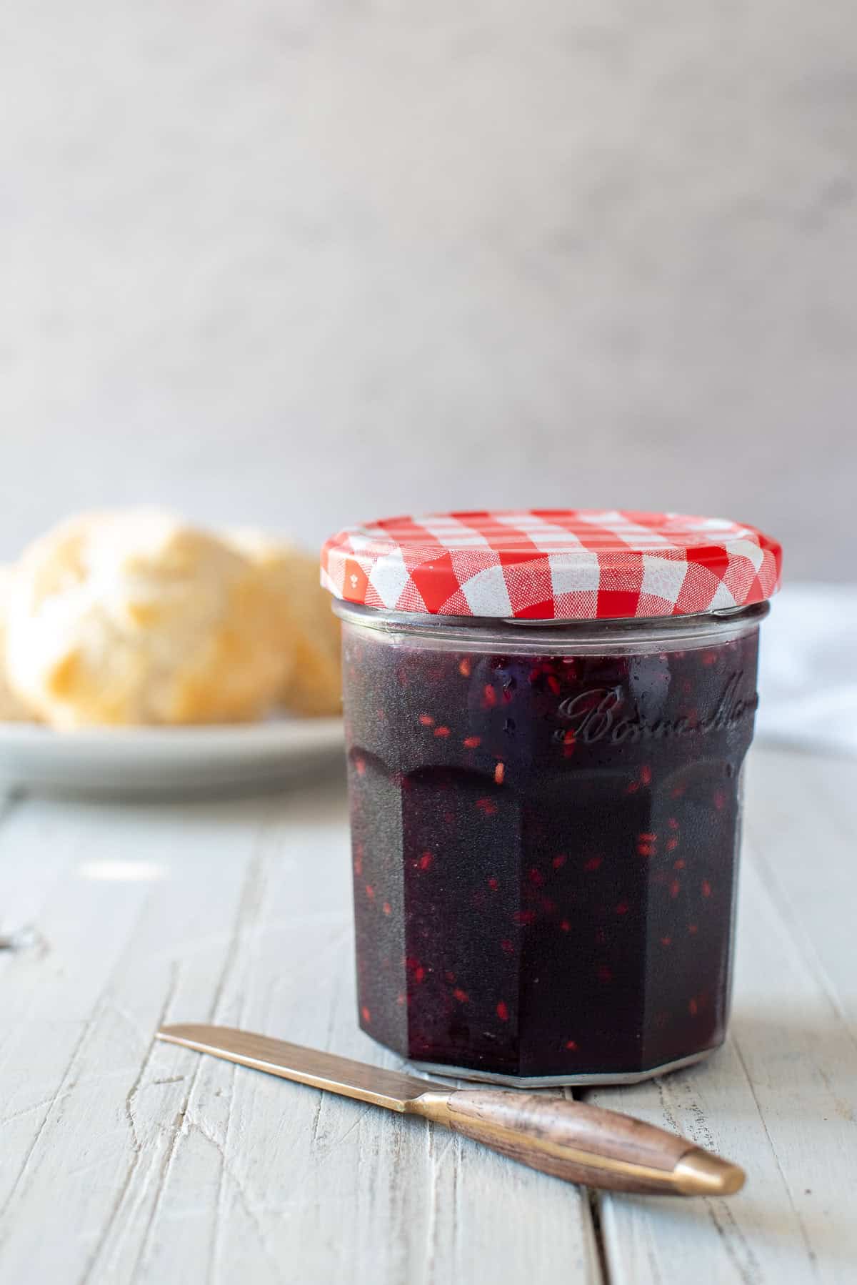 blackberry jam in a glass jar with a red and white checkered lid. A knife is laying on the white table and biscuits are seen in the background.