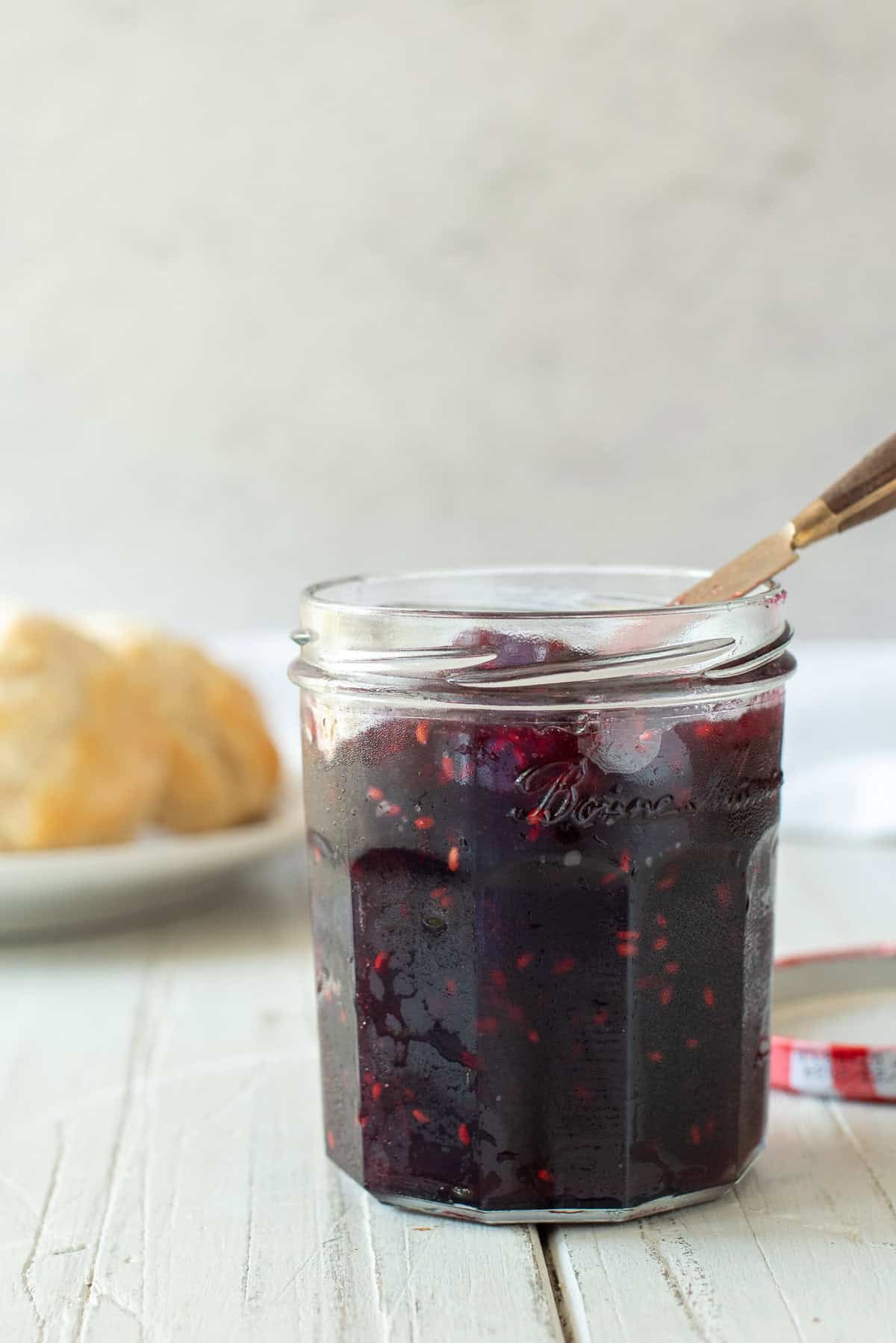blackberry freezer jam in a glass jar with a knife stuck in it. Biscuits are on a plate in the background.