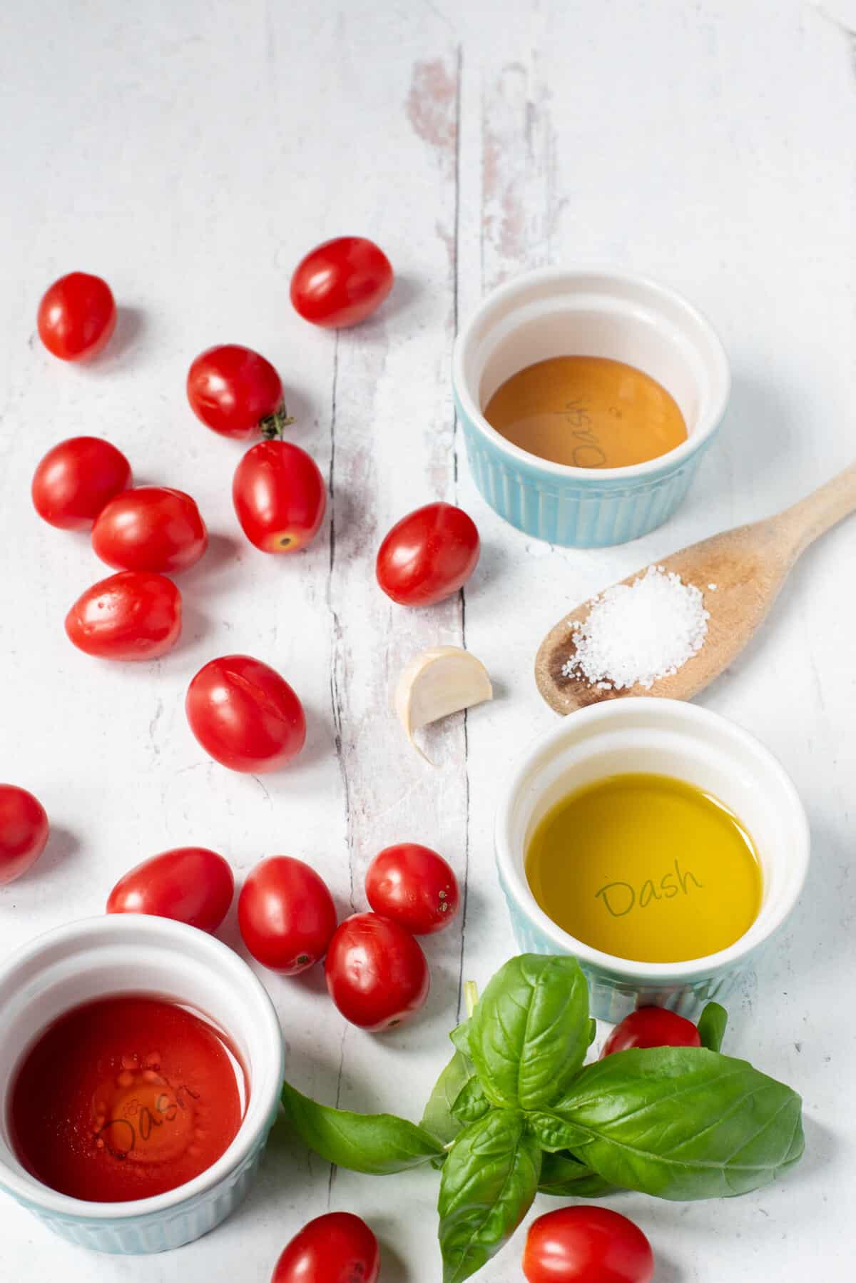 ingredients needed for marinated tomatoes: grape tomatoes, garlic, basil, a wooden spoon of Kosher salt, and small bowls of honey, olive oil, and red wine vinegar on a white background.