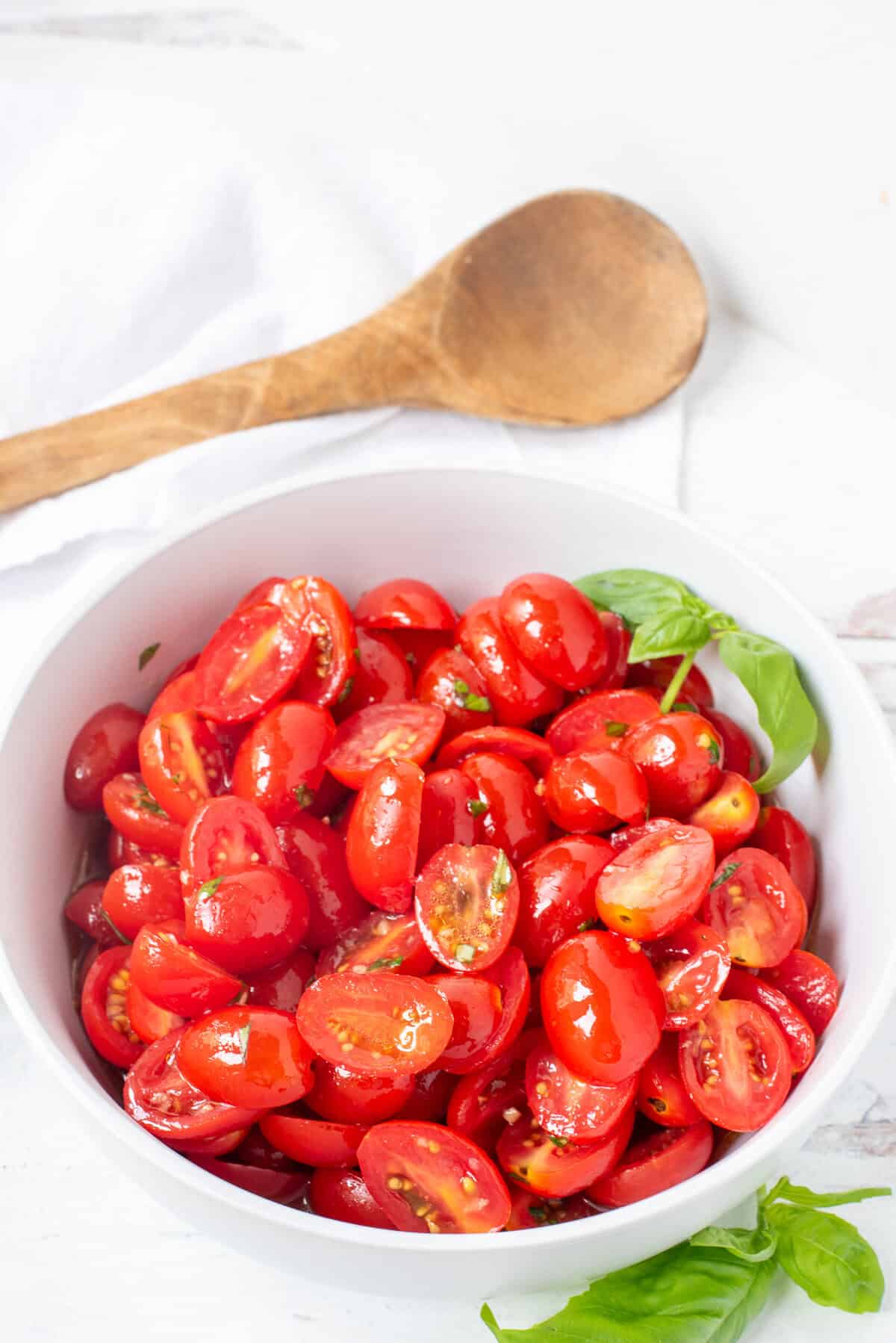 white bowl of marinated tomatoes on a white background with a wooden spoon laying nearby