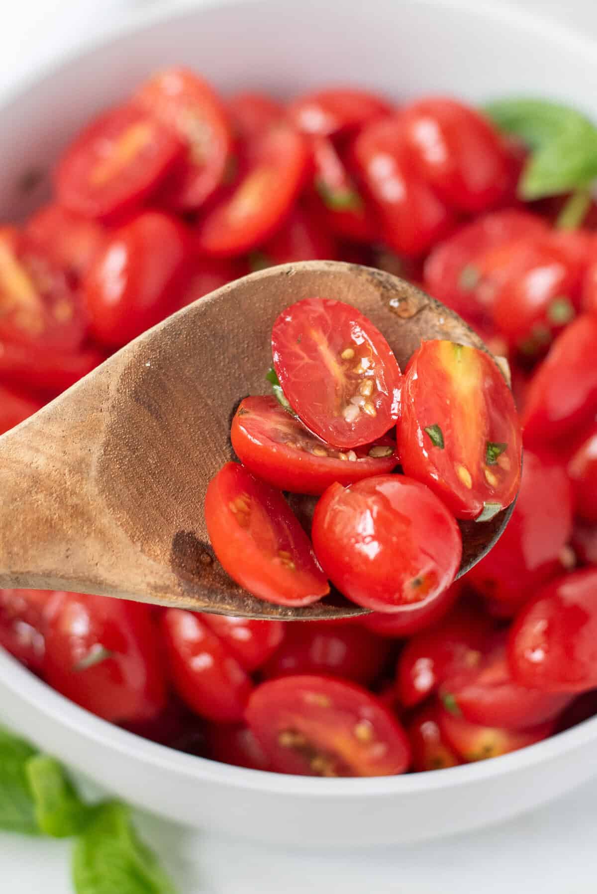 Wooden spoon filled with marinated tomatoes with a bowl of marinated tomatoes in the background.