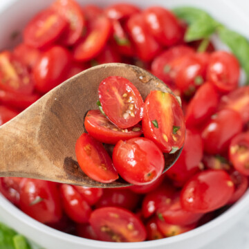 marinated tomatoes in a wooden spoon with a bowl of tomatoes in the background.