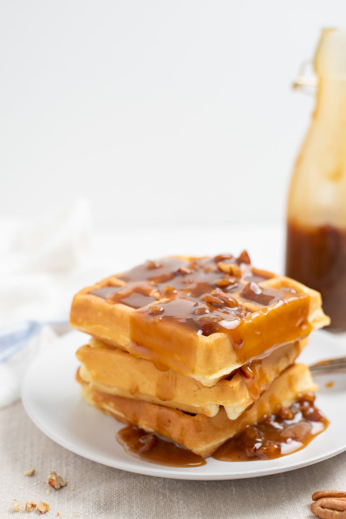 A stack of belgian waffles on a white plate topped with syrup and pecans