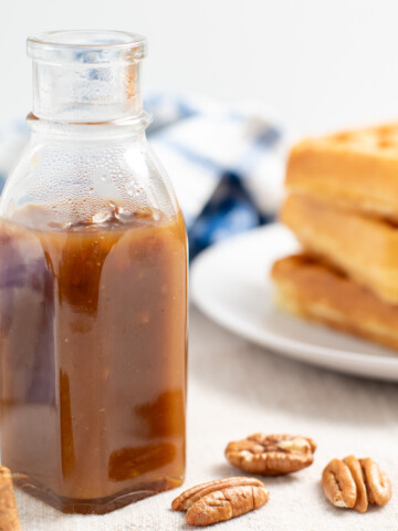 a glass jar of butter pecan syrup surrounded by pecans with a plate of belgian waffles in the background