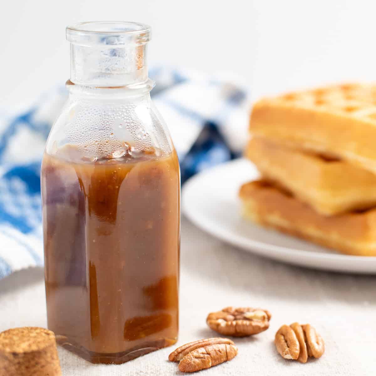 a glass jar of butter pecan syrup surrounded by pecans with a plate of belgian waffles in the background