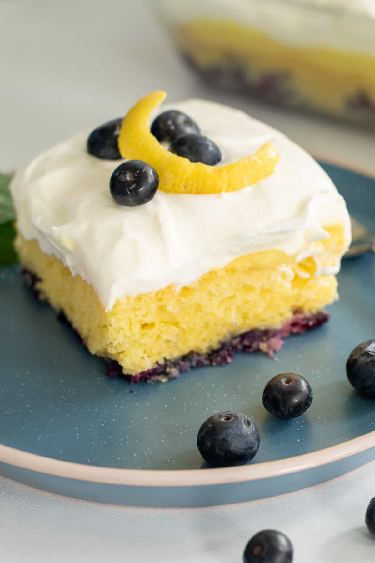 lemon blueberry poke cake topped with fresh berries and a lemon curl on a blue plate
