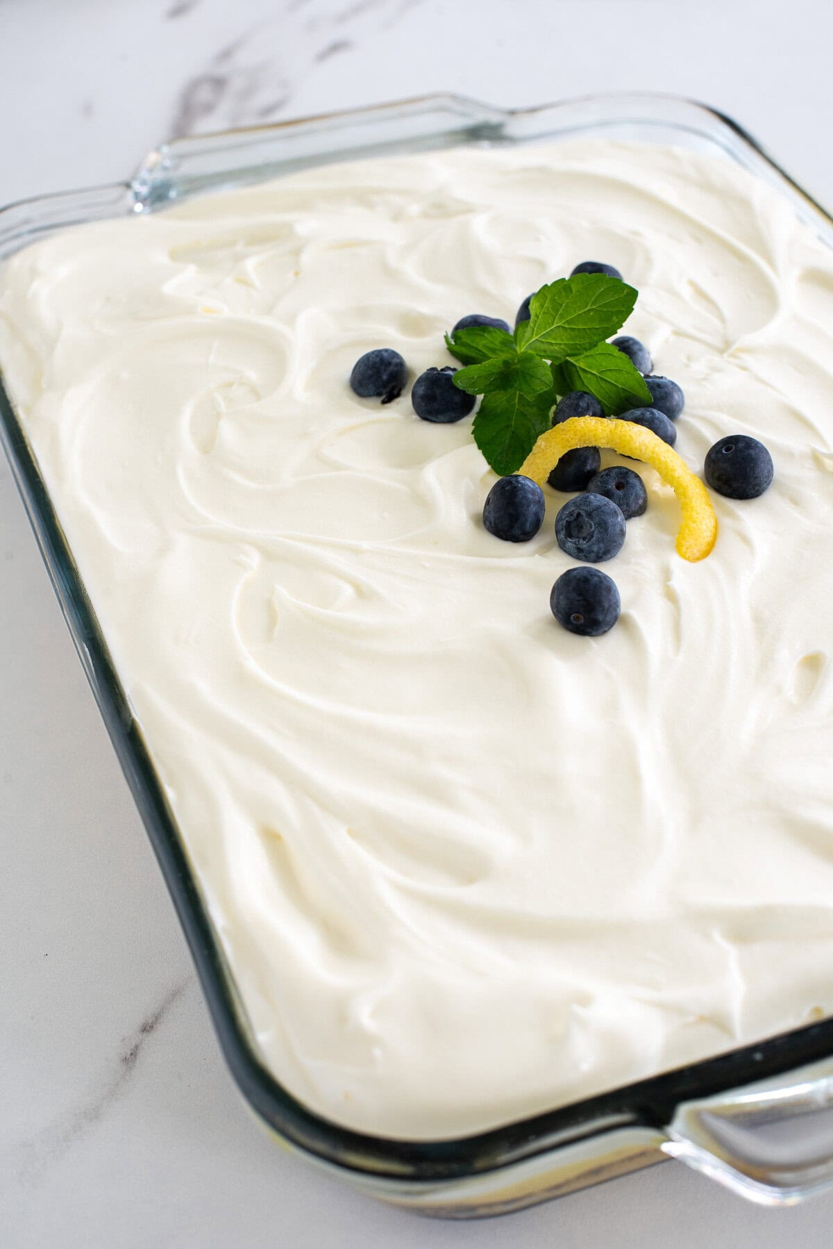 A whole lemon blueberry poke cake topped with whipped cream cheese topping and fresh blueberries