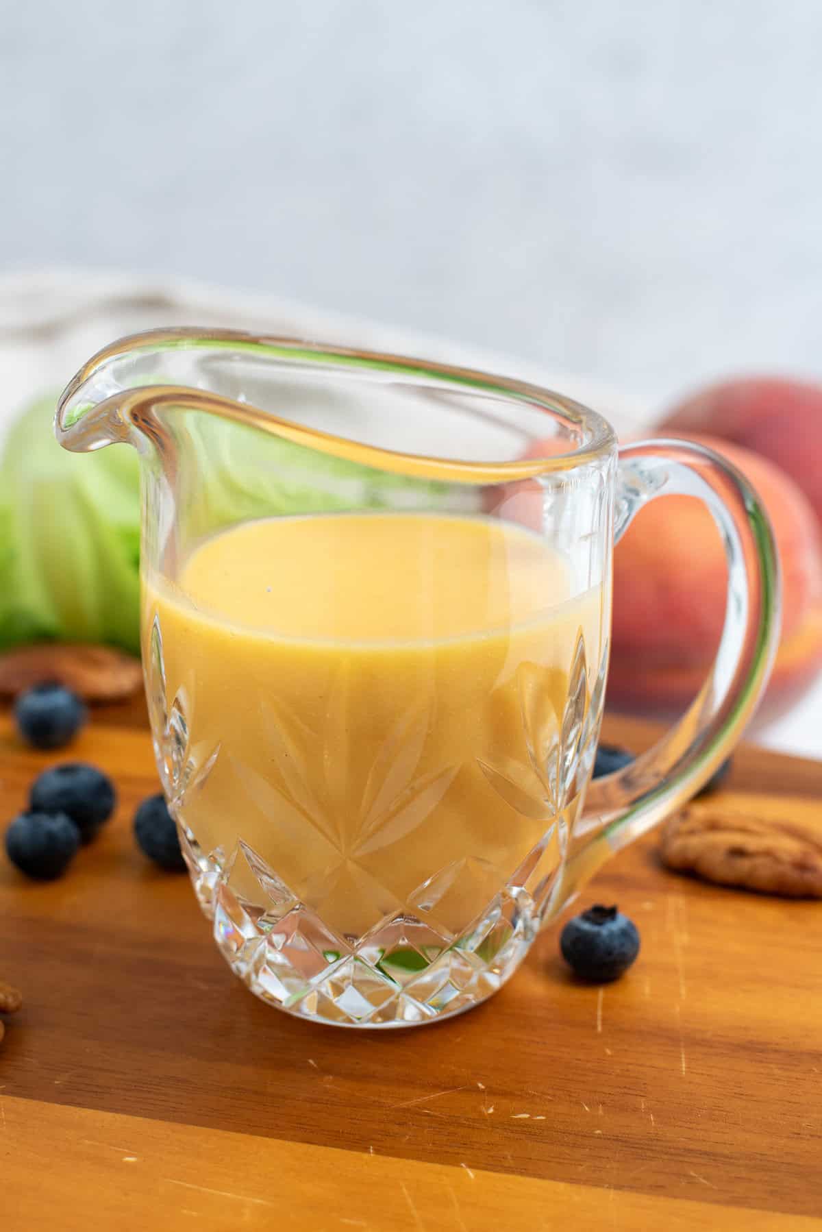 peach vinaigrette in a glass pitcher on a wooden cutting board with fruits and vegetables around it