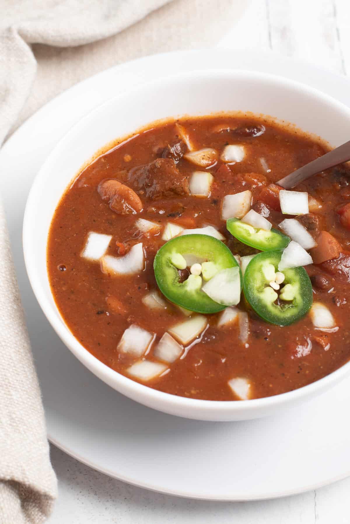 A bowl of chili topped with diced onions and sliced jalapeno peppers. 