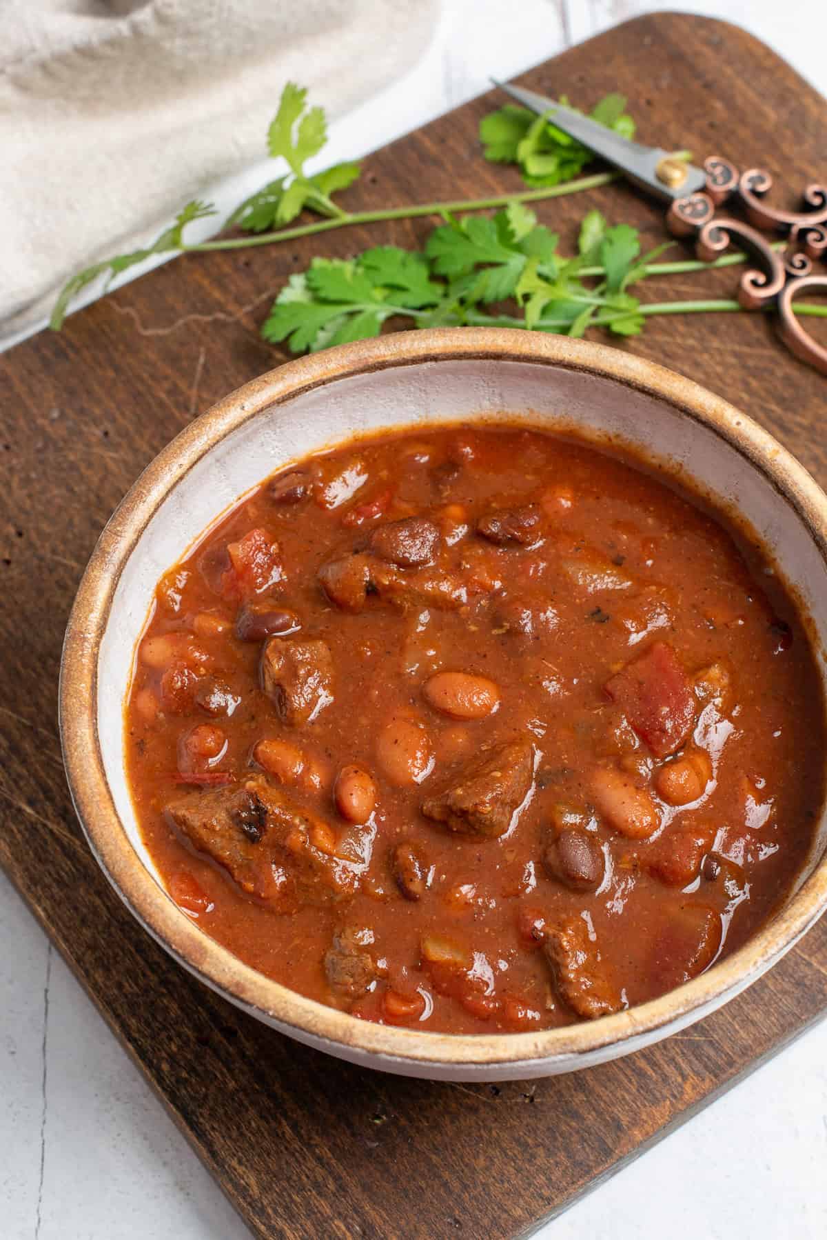 A bowl of chuck roast chili on a wooden board with cilantro in background.