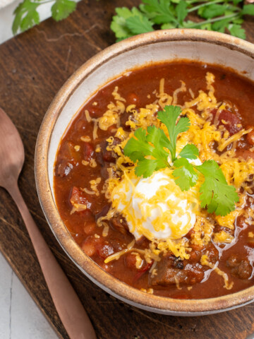 chuck roast chili on a wooden board with a spoon, topped with sour cream, cheese, and green onions.