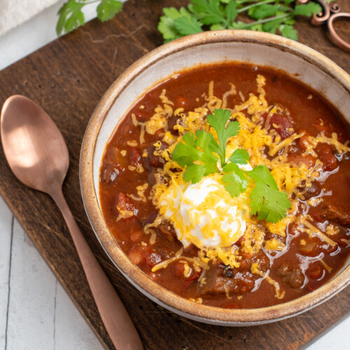 chuck roast chili on a wooden board with a spoon, topped with sour cream, cheese, and green onions.