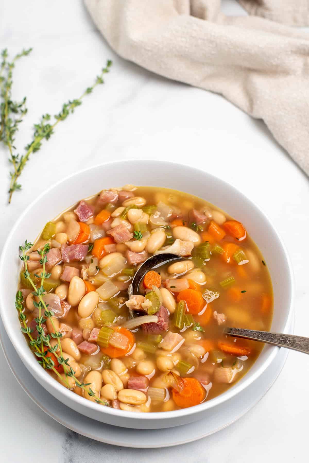 ham soup with beans in a  white bowl with a spoon and fresh thyme. 