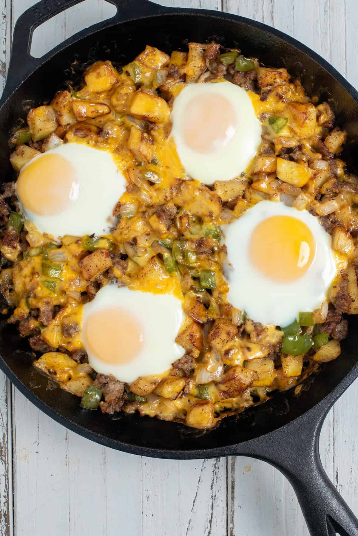 A cast iron skillet filled with potatoes, sausage, peppers, onions, and cheddar cheese, topped with a fried egg.