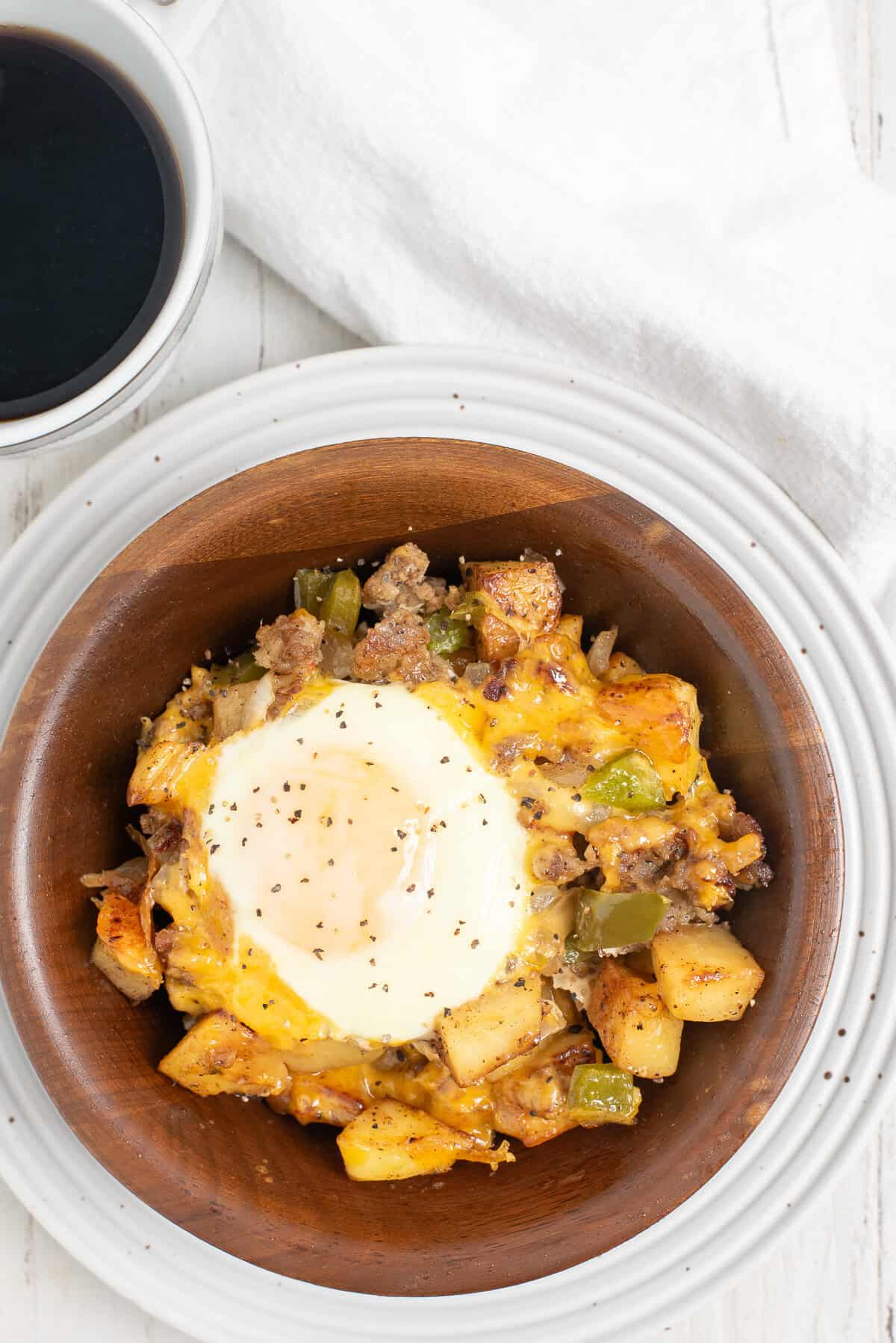 A brown bowl full of sausages, potatoes, peppers, onions, and cheddar cheese, topped with a seasoned fried egg.