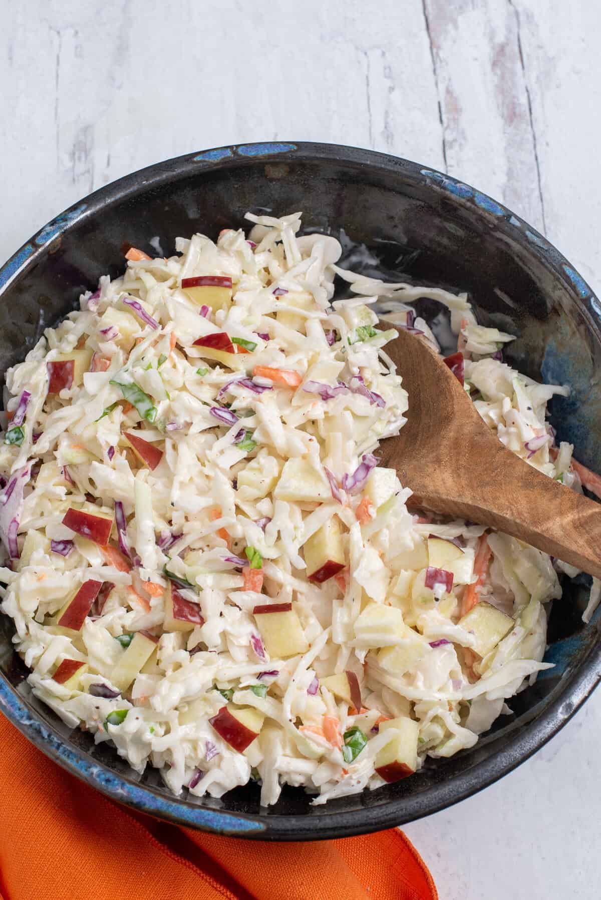 A dark bowl full of cabbage and apple salad with a wooden spoon and an orange napkin.