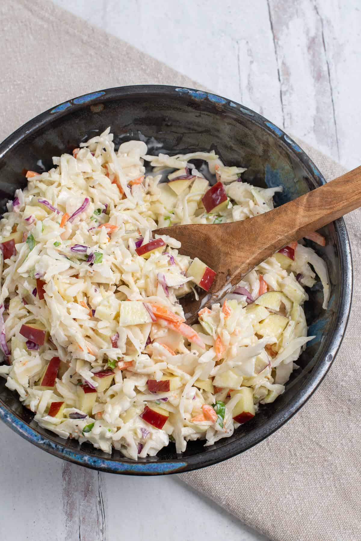 A dark bowl full of apple and cabbage slaw with a wooden spoon and a linen napkin.