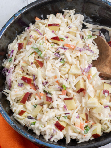 A dark bowl full of cabbage and apple slaw with a wooden spoon and an orange napkin.