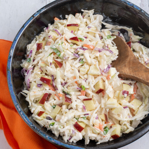 A dark bowl full of cabbage and apple slaw with a wooden spoon and an orange napkin.