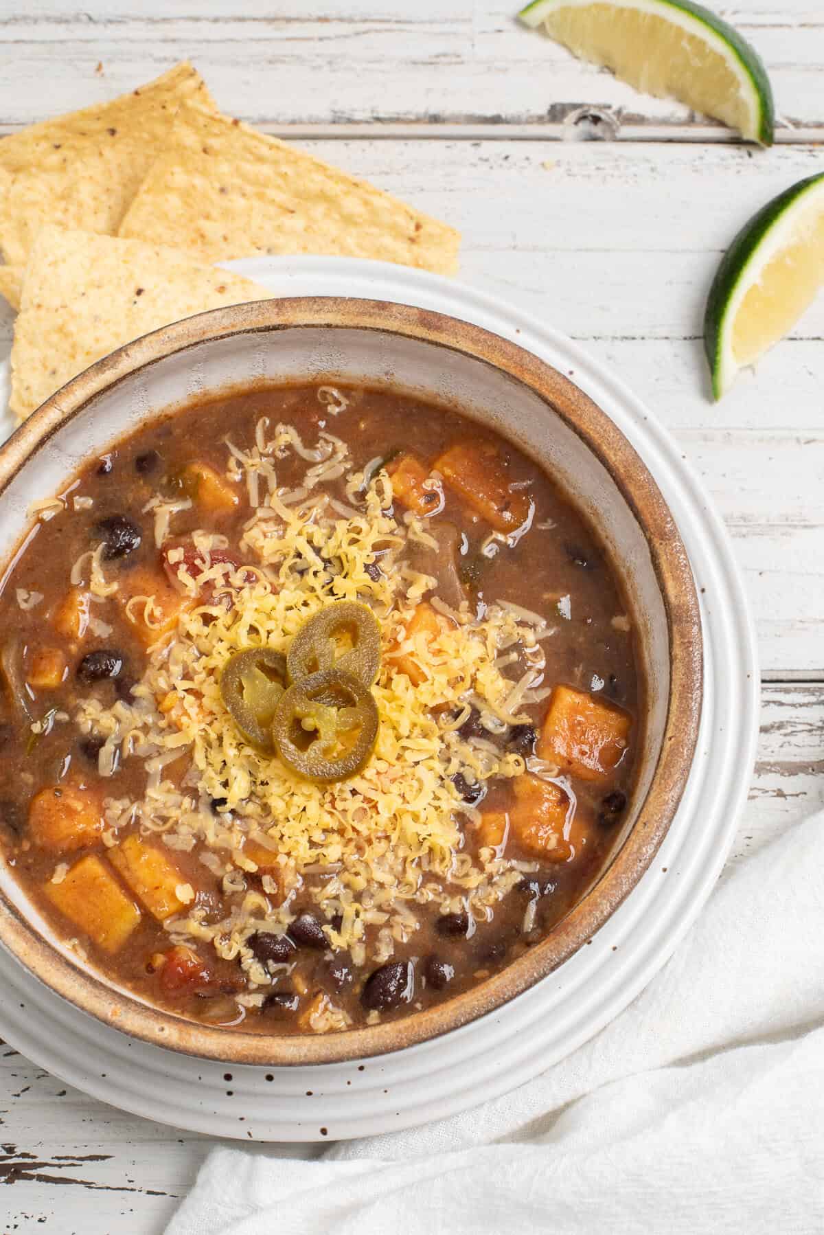 Black bean sweet potato soup in a tan bowl topped with cheese and sliced jalapenos with tortilla chips and a lime wedge nearby.