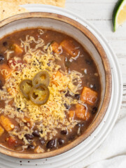 Black bean sweet potato soup in a tan bowl topped with cheese and sliced jalapenos with tortilla chips and a lime wedge nearby.
