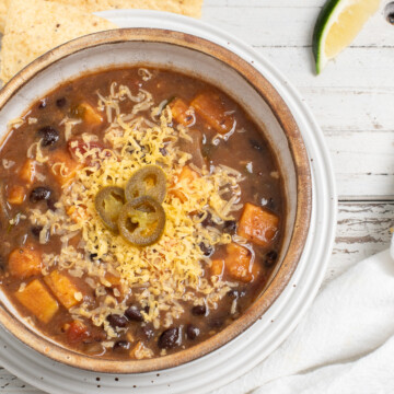 Black bean sweet potato soup in a tan bowl topped with cheese and sliced jalapenos with tortilla chips and a lime wedge nearby.